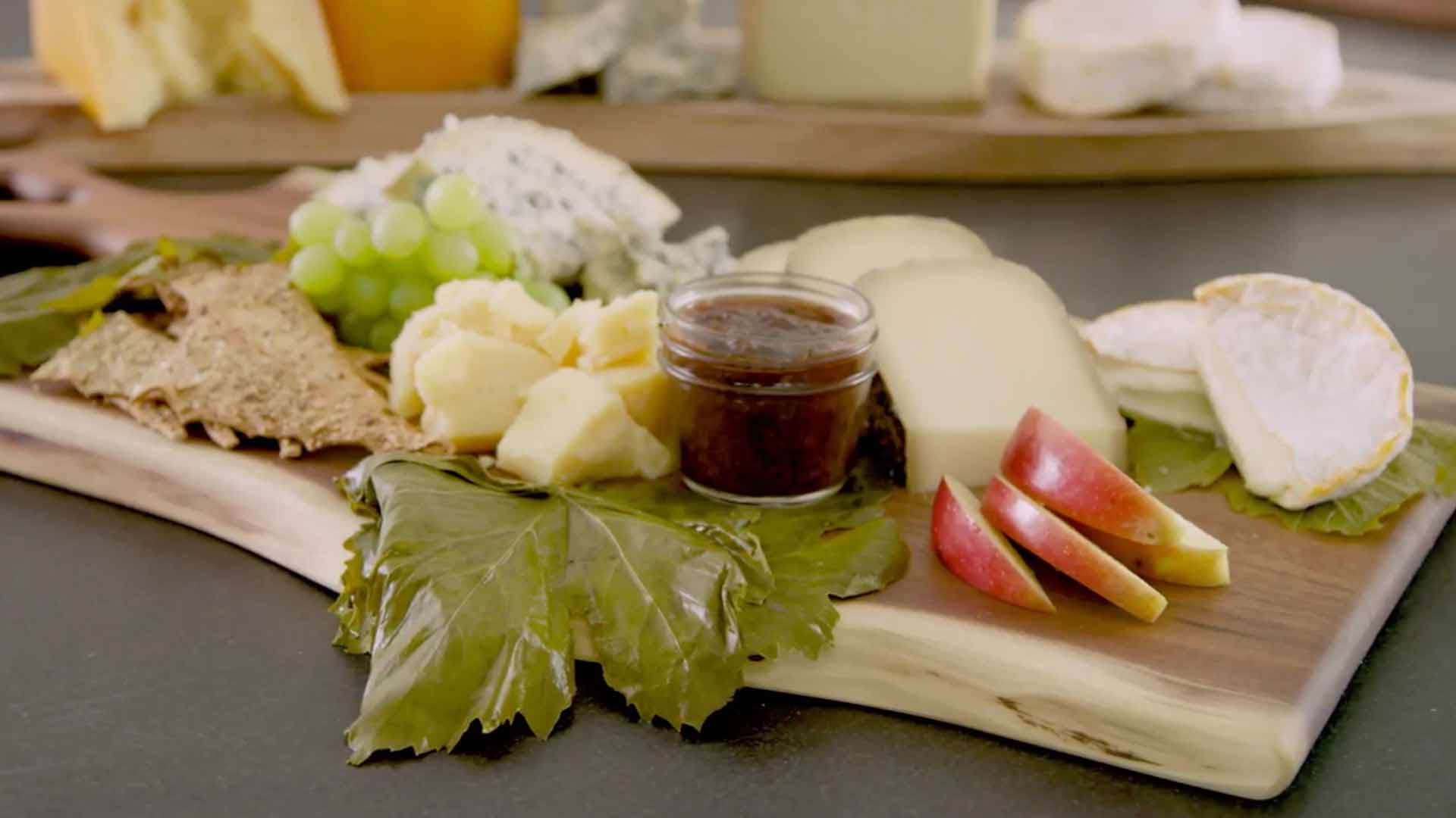 Cheese wedges next to small jar of jam, grapes apple slices on wooden serving board. 