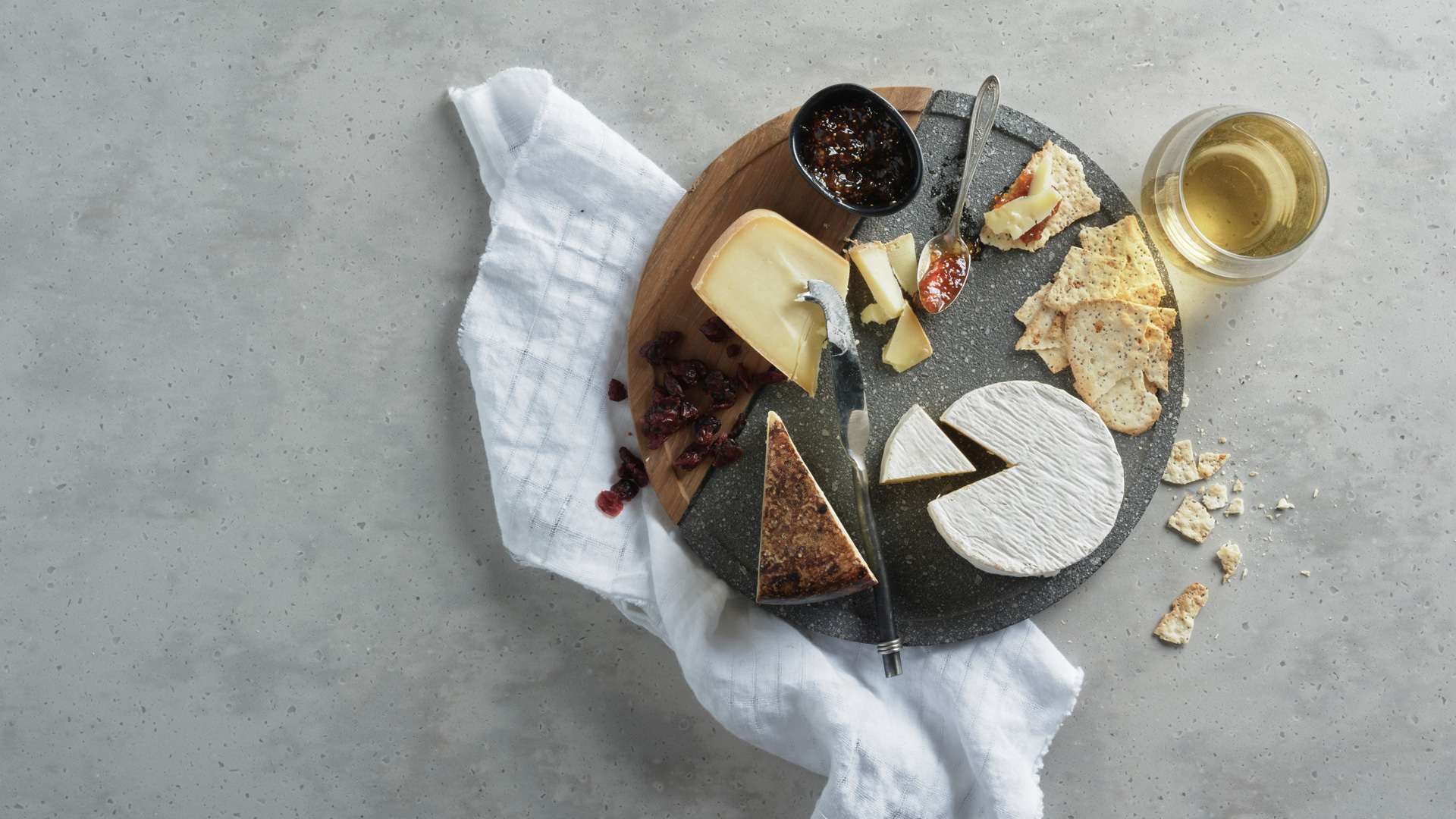 Selection of three cheeses and wine on a sphere grey marble board, with scattered cranberries, crackers, and jam spread in a small bowl with a white napkin on the left side. 