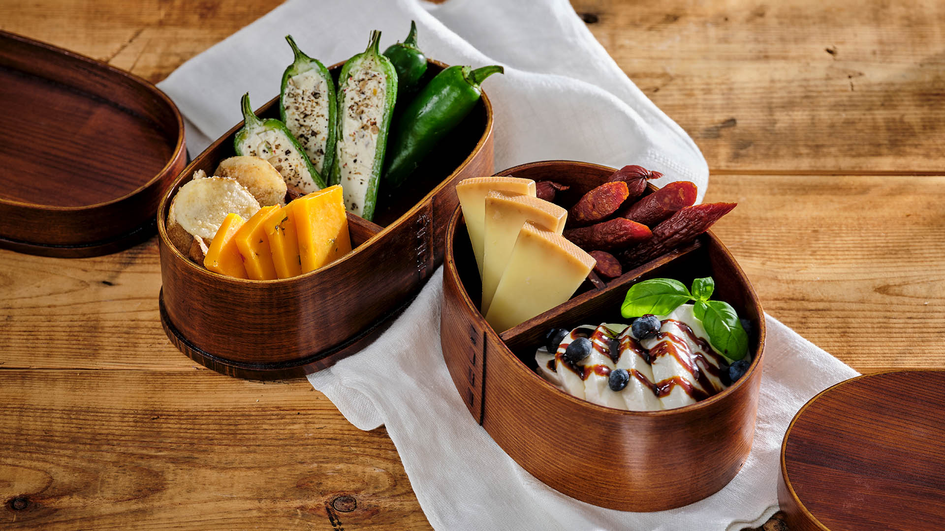 Two wooden bento boxes containing various types of artisanal cheese, fruits, and vegetables. 