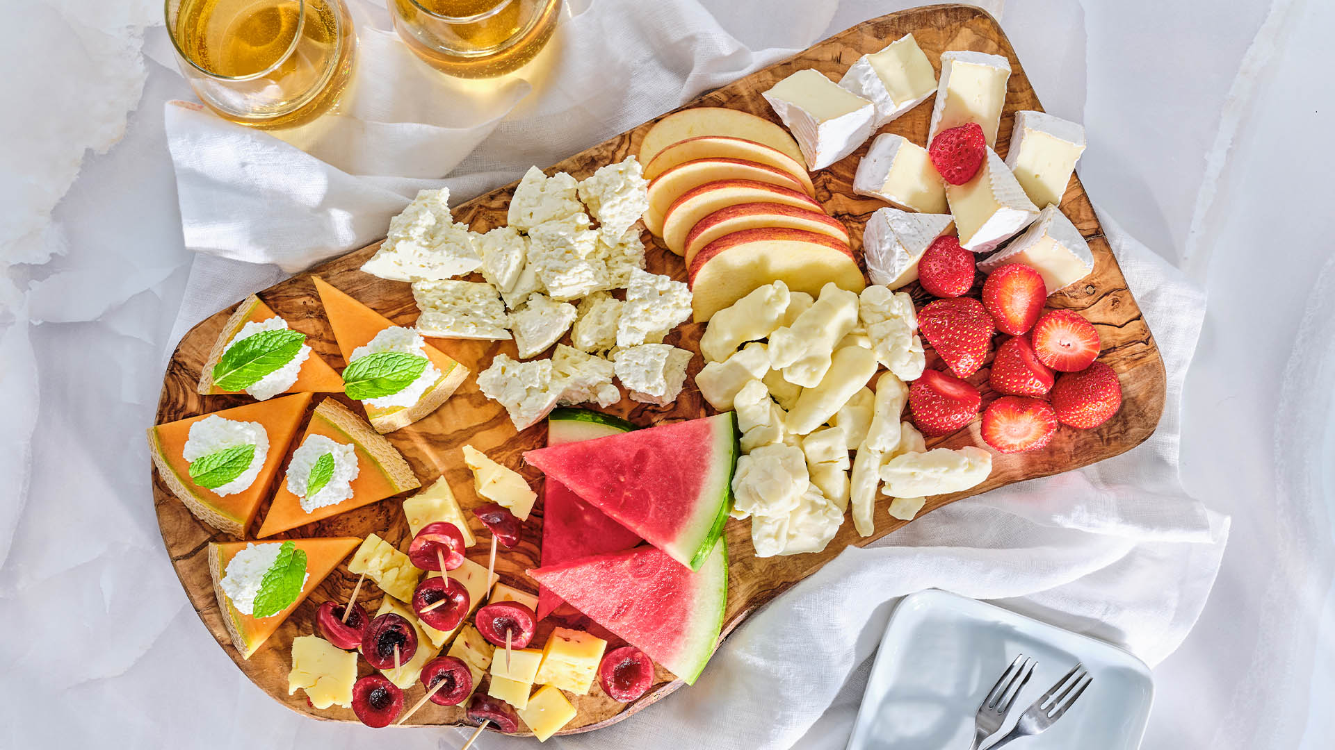 A wooden board on a marble surface containing various types of artisanal cheeses and in season summer fruits.