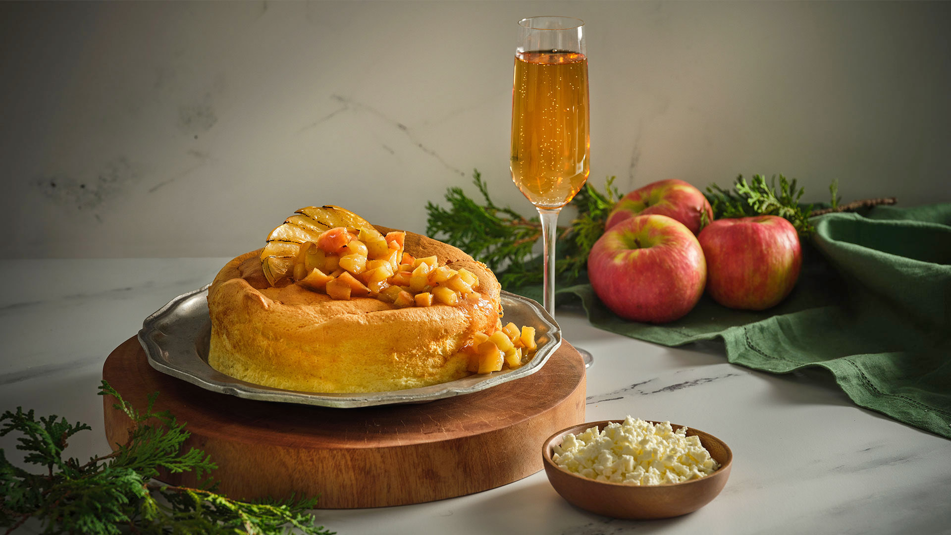 A Feta Cheesecake with Apple Compote on a round silver platter on top of a round wooden serving board next to a tall glass of wine next to 3 apples and a green napkin.
